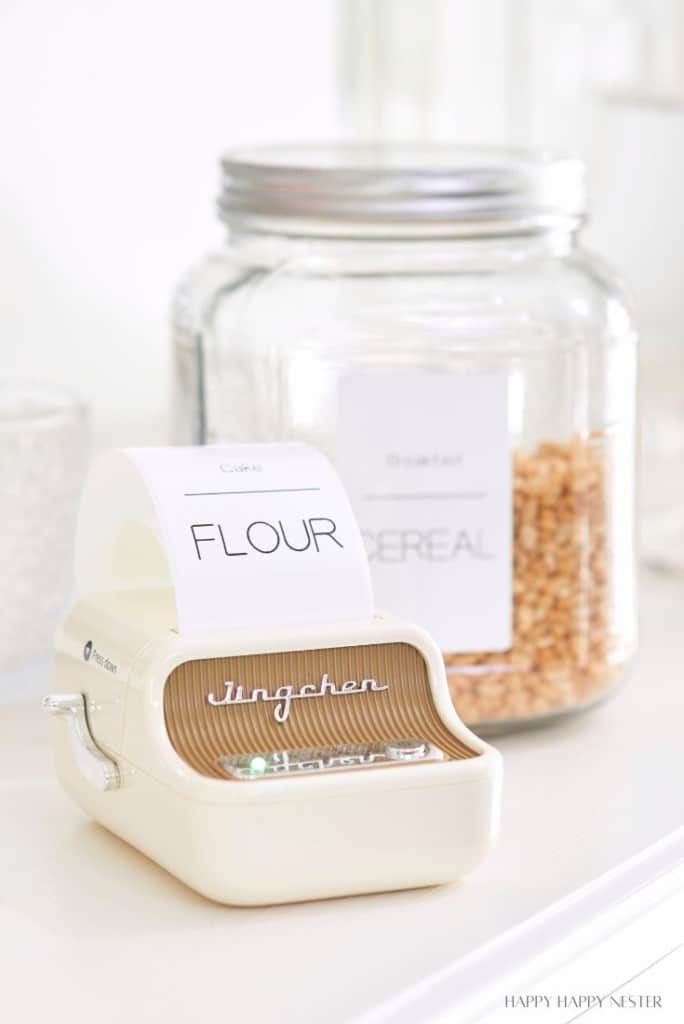 A vintage label maker rests on a white surface, printing "FLOUR" as a glass jar labeled "CEREAL" stands behind it. This scene perfectly captures the essence of organizing a small pantry, enhancing both style and efficiency in your kitchen space.