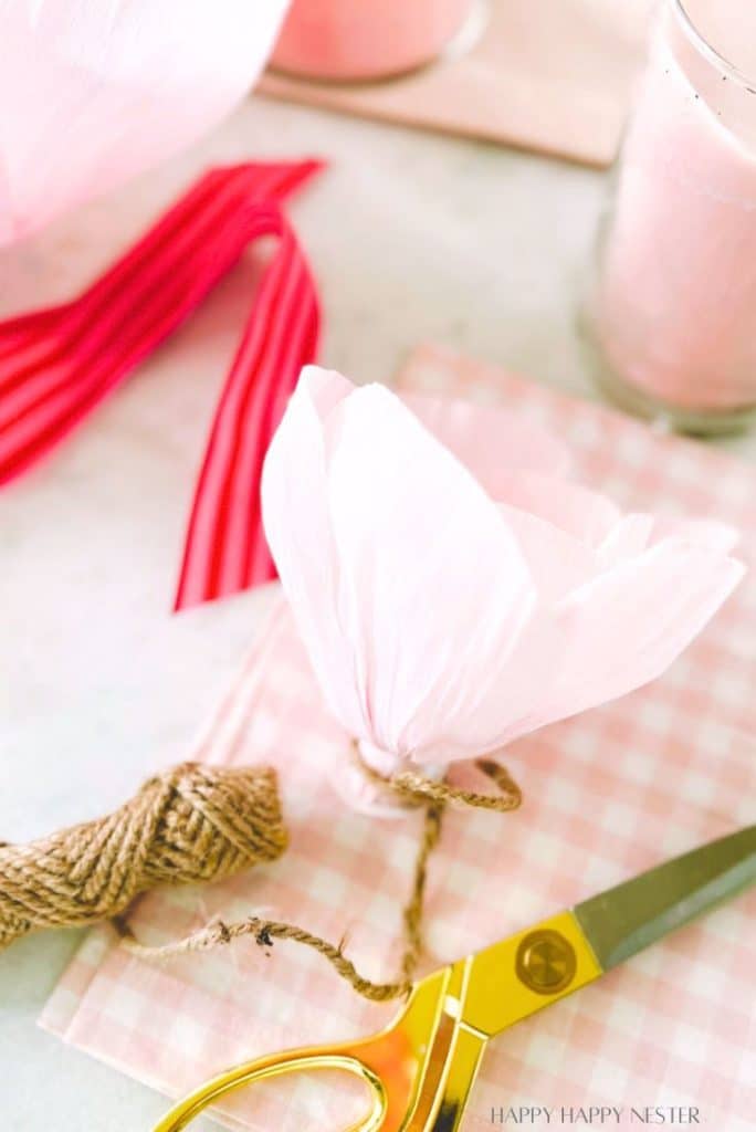 A partially assembled pink crepe paper flower rests on a pink checkered cloth, perfect for a DIY garland project. Nearby are gold scissors, twine, and red crepe paper strips. A glass with a pink drink and a pink candle enhance the charming scene in the background.