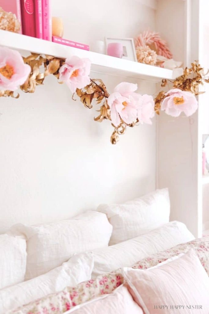 A cozy bedroom features white bedding with floral patterns. Above the bed, a white shelf showcases pink books and decor, with a garland of pink and gold flowers—just like my favorite spring crafts—hanging underneath. The wall is a soft white, enhancing the serene atmosphere.