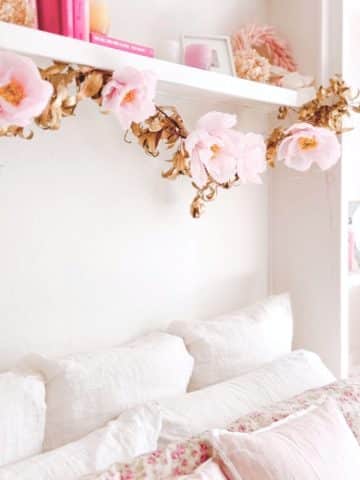 Cozy bedroom with a white bed adorned with a floral-patterned blanket. A DIY flower garland drapes gracefully alongside pink flowers and golden leaves on the shelf above. Pink books and decorative items are neatly arranged, enhancing the charm of this serene space.