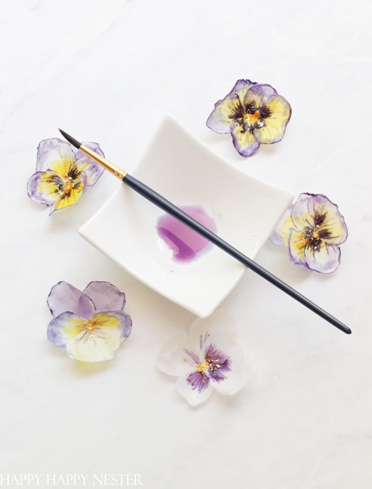 A paintbrush with a purple tip rests on a white, square dish containing a small amount of purple paint. Surrounding the dish are delicate watercolor flowers in shades of purple and yellow, perfect for easy flower paper crafts, all beautifully arranged on a white surface.