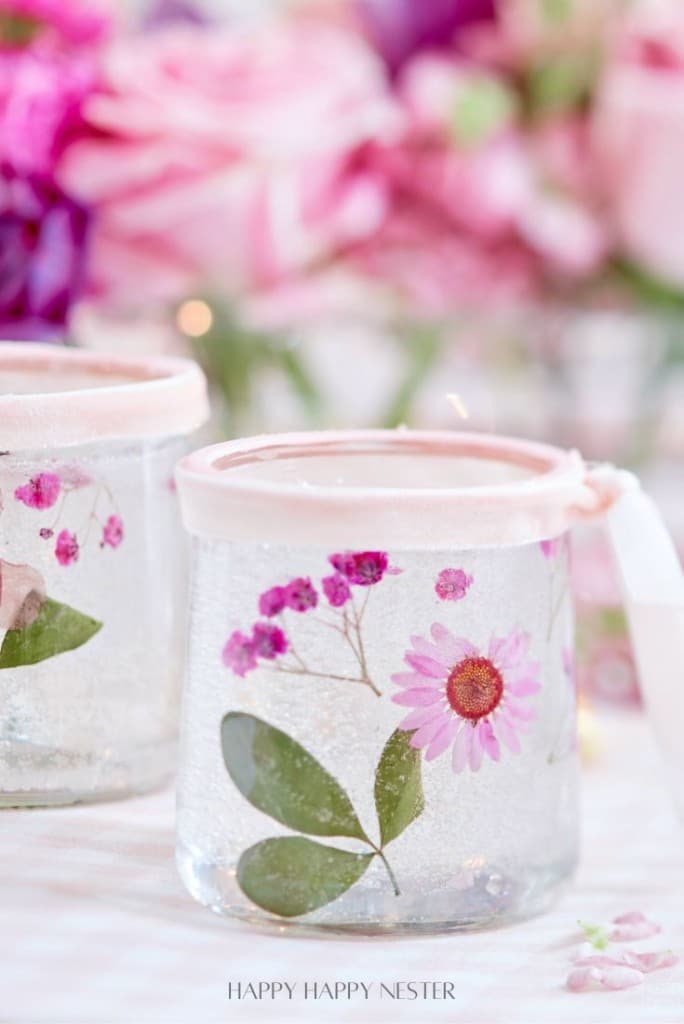 Two glass jars, perfect for making candles with dried flowers, are adorned with pressed pink flowers and green leaves, tied with pink ribbons. In the background, blurred pink and purple flowers add to their soft, elegant appearance.