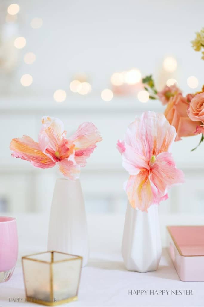 Two white vases holding large pink and orange flowers sit on a table. Next to them, a pink candle and a gold geometric holder complete the scene, enhanced by soft-focus lights for a cozy ambiance. It's as delightful as easy flower paper crafts for brightening any room.