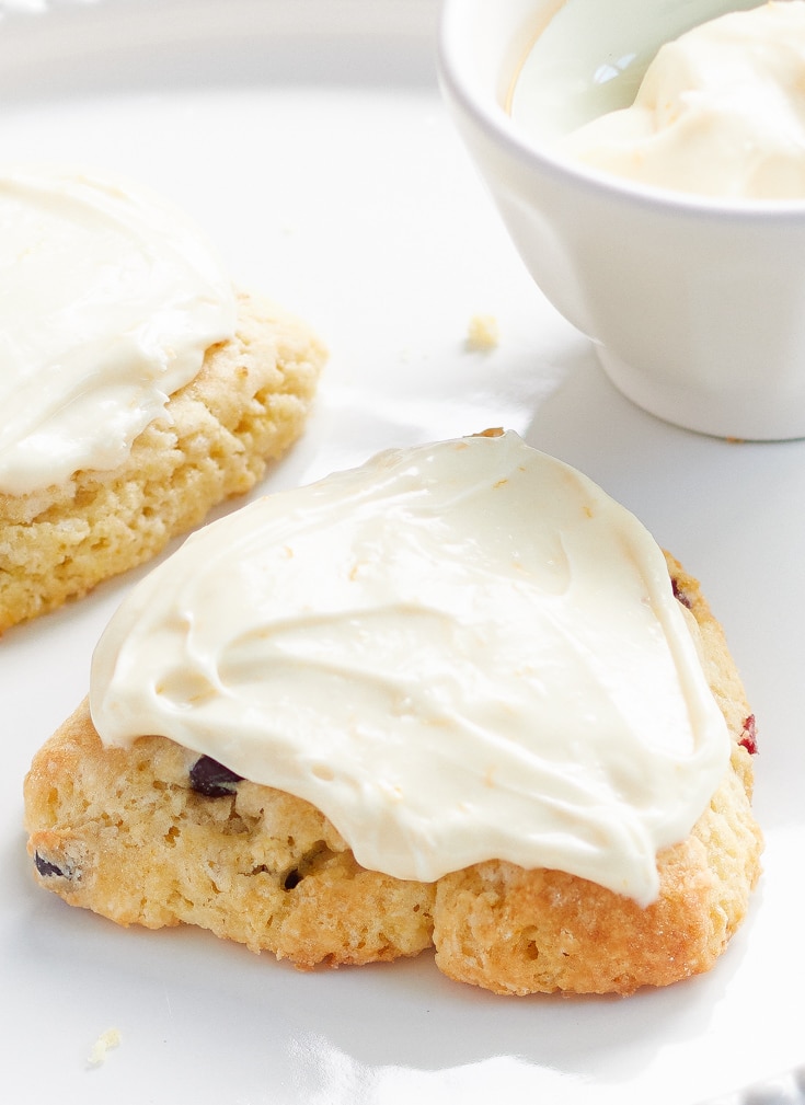 A scone, one of my favorite desserts, is topped with creamy white frosting on a white plate. Another partially visible scone and a small bowl of cream linger in the background.