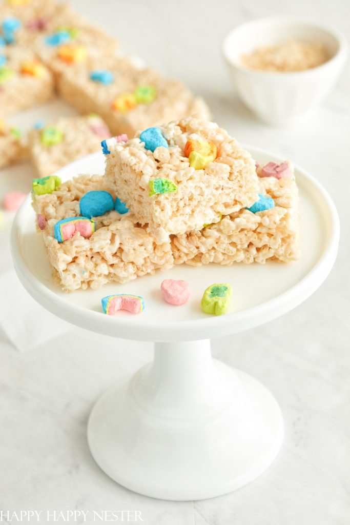 Stacked on a white cake stand, the rice crispy treats with colorful marshmallows sparkle like gems. Behind them, more treats and a bowl of cereal await on the marble surface, embodying all the joy of my favorite desserts.