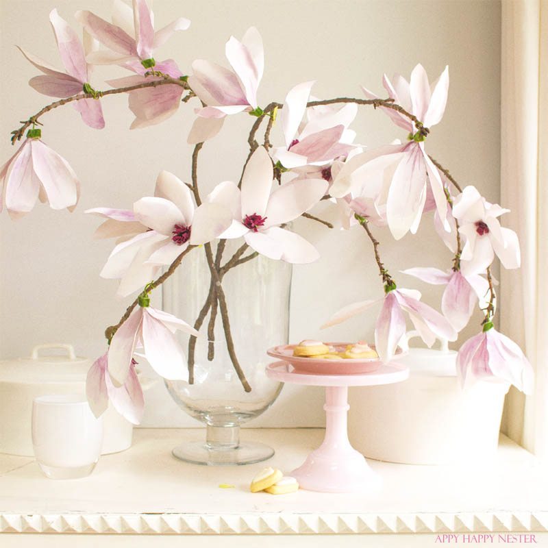 Vase with pink magnolia branches on a white table, complemented by easy flower paper crafts. A pink cake stand nearby holds macarons, while soft lighting creates a serene atmosphere.