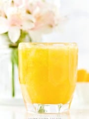 A glass of orange juice takes center stage, filled to the brim with a frothy top, resembling an easy summer mocktail recipe. In the background, a blurred bouquet of pink and white flowers in a glass jar adds a touch of elegance to this bright and inviting scene.