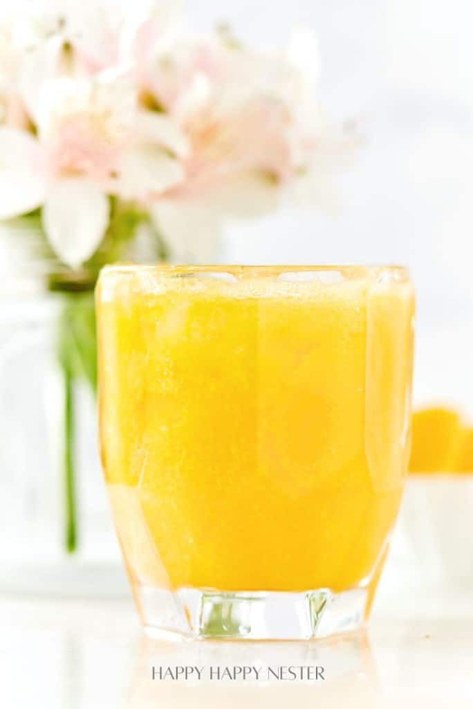 A glass of orange juice takes center stage, filled to the brim with a frothy top, resembling an easy summer mocktail recipe. In the background, a blurred bouquet of pink and white flowers in a glass jar adds a touch of elegance to this bright and inviting scene.