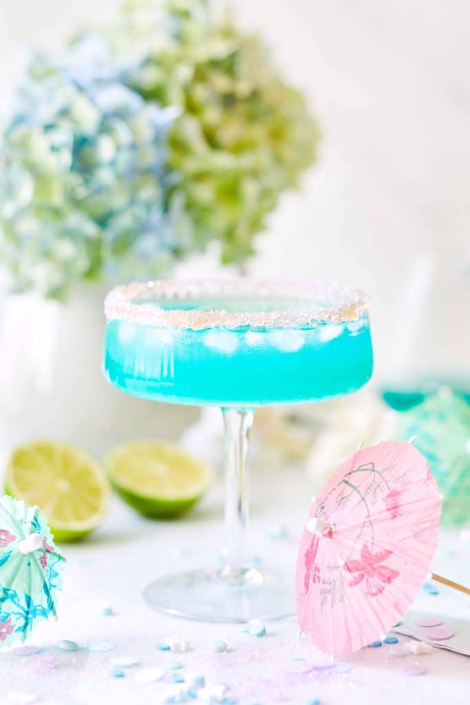 A vibrant blue cocktail in a glass with a sugared rim, garnished with a pink paper umbrella, ranks among the best summer drinks. The backdrop of hydrangeas and halved limes enhances its allure, while decorative umbrellas and petals are scattered around the glass.