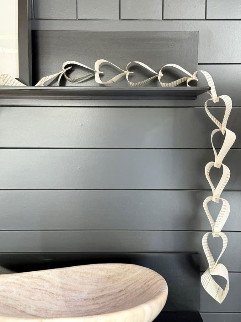 A decorative paper chain of heart shapes is draped over a dark wooden shelf. The wall behind is made of horizontal wooden panels. A light-colored, stone-textured bowl, accompanied by candles with dried flowers, is placed on the counter below the shelf.