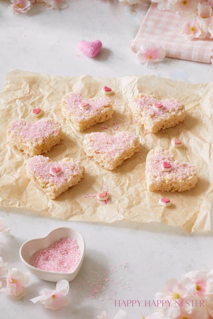 Heart-shaped rice crispy treats with pink sprinkles are arranged on parchment paper, embodying my favorite desserts. Nearby, a small heart-shaped bowl overflows with pink sprinkles. Decor accents include pink hearts and flowers on a light background, adding to the charm.