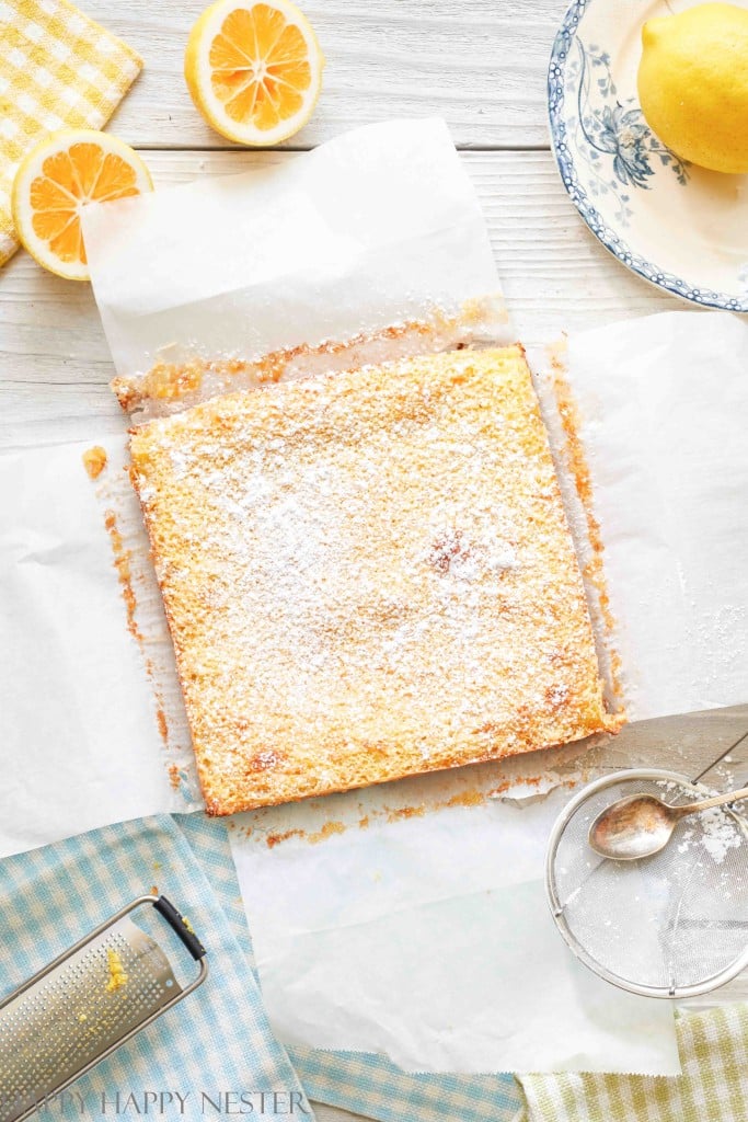 A tray of freshly baked lemon bars, one of my favorite desserts, is dusted with powdered sugar on parchment paper. Surrounded by halved lemons, a lemon squeezer, and a decorative plate on a white wooden table with checkered cloths.