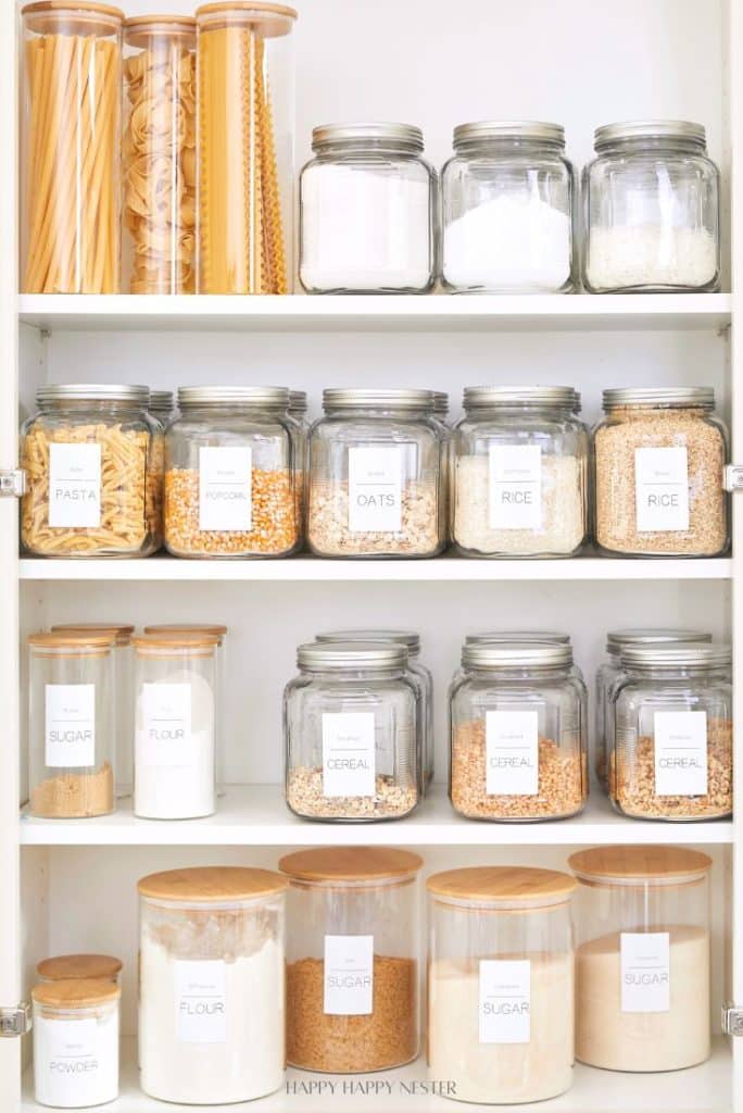 A small pantry boasts a neatly organized shelf adorned with glass jars filled with dry foods like pasta, popcorn, and oats. Each jar, crowned with a wooden lid and marked by a white label, lines up in perfect rows, maximizing space while maintaining an aesthetic allure.