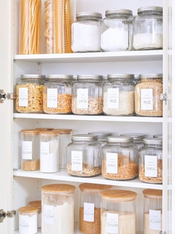 Organizing a small pantry is made simple with a neatly arranged shelf of glass jars containing pasta, oats, rice, sugar, flour, and cereal. Each jar features a wooden lid and clear label, perfectly aligned across three shelves for effortless access and visual appeal.