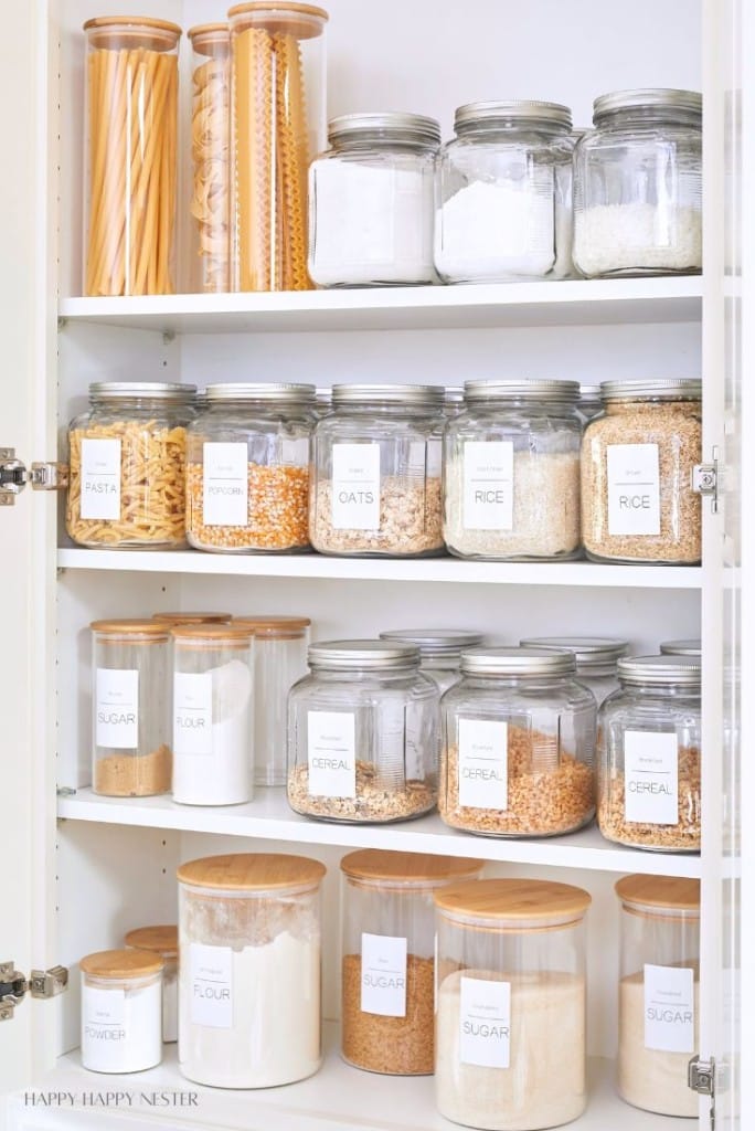 Organizing a small pantry is made simple with a neatly arranged shelf of glass jars containing pasta, oats, rice, sugar, flour, and cereal. Each jar features a wooden lid and clear label, perfectly aligned across three shelves for effortless access and visual appeal.