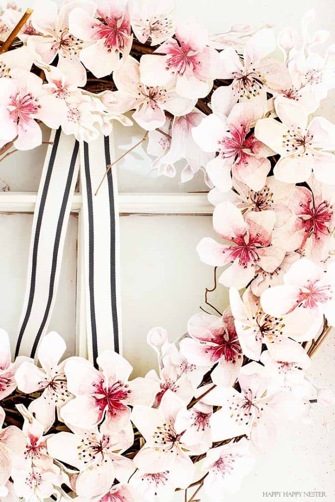 A floral wreath made of pink and white blossoms with dark centers elegantly hangs on a door, adorned with a black and white striped ribbon. Its delicate petals embody the charm of easy flower paper crafts, adding an artistic touch to any entrance.