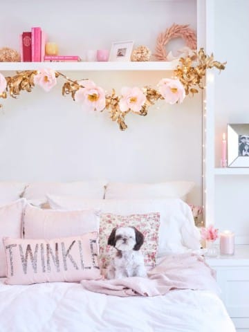 A cozy bedroom features a white bed with decorative pillows, one labeled "TWINKLE." A small black and white dog sits on the bed. Above, a DIY flower garland adds charm to the shelf adorned with pink and gold flowers, books, and framed photos. Soft lighting adds a warm touch.