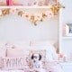 A cozy bedroom features a white bed with decorative pillows, one labeled "TWINKLE." A small black and white dog sits on the bed. Above, a DIY flower garland adds charm to the shelf adorned with pink and gold flowers, books, and framed photos. Soft lighting adds a warm touch.