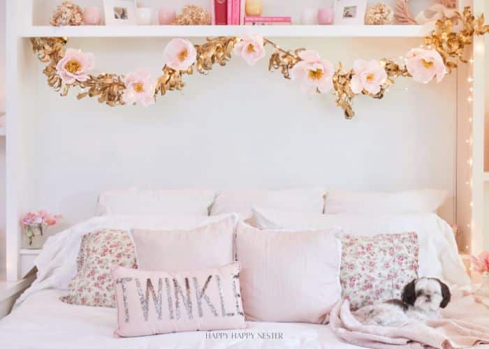 A cozy bedroom features a bed with white bedding and floral-patterned pillows. A decorative "Twinkle" pillow sits in front. Above the bed, a garland of pink and gold flowers is draped. A small dog rests on the bed. Shelves display small items.