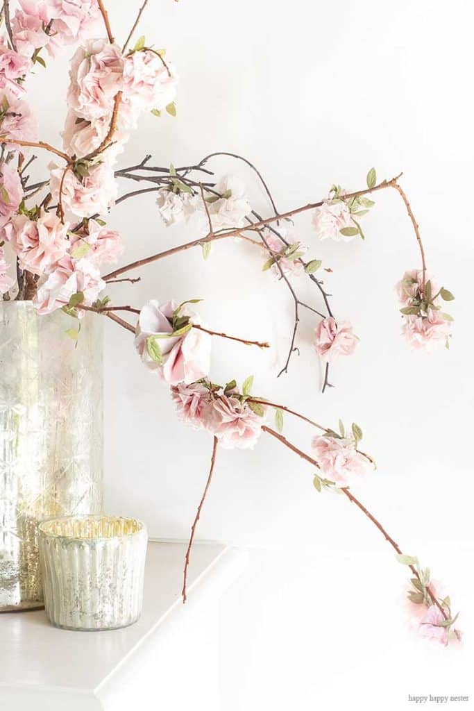 A glass vase holds branches adorned with soft pink and white cherry blossoms. The branches extend gracefully, displaying delicate flowers and small green leaves against a plain white background. A smaller textured glass candle holder sits beside the vase, reminiscent of easy flower paper crafts in its simplicity.