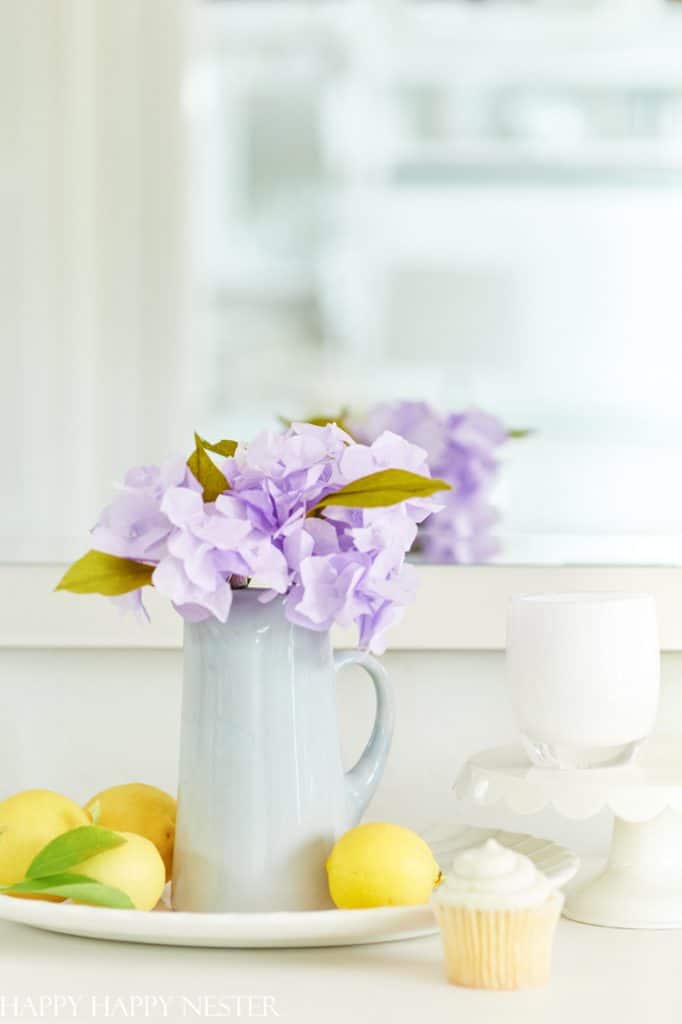 A light blue vase with purple flowers, beside three yellow lemons and a green leaf. A frosted cupcake sits on a white cake stand near a white cup. The background features a mirror reflecting the setup, perfect inspiration for easy flower paper crafts.