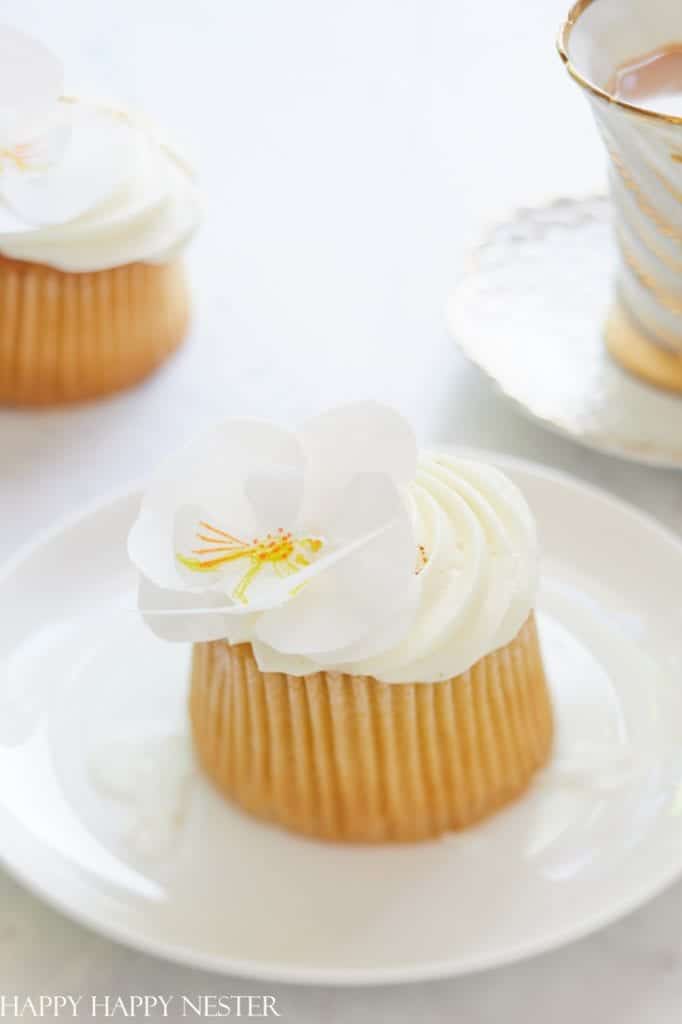 A small cake with white frosting is topped by a delicate flower garnish that mimics easy flower paper crafts. The cake rests elegantly on a white plate, while in the background, an ornate teacup with a gold design sits partially visible on its matching saucer.