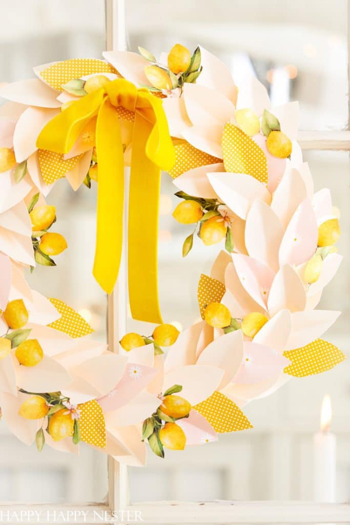 A decorative wreath made of easy flower paper crafts like magnolia blooms and lemons hangs on a window. It features a large, bright yellow bow at the top and has a soft, pastel color scheme with hints of green and yellow.