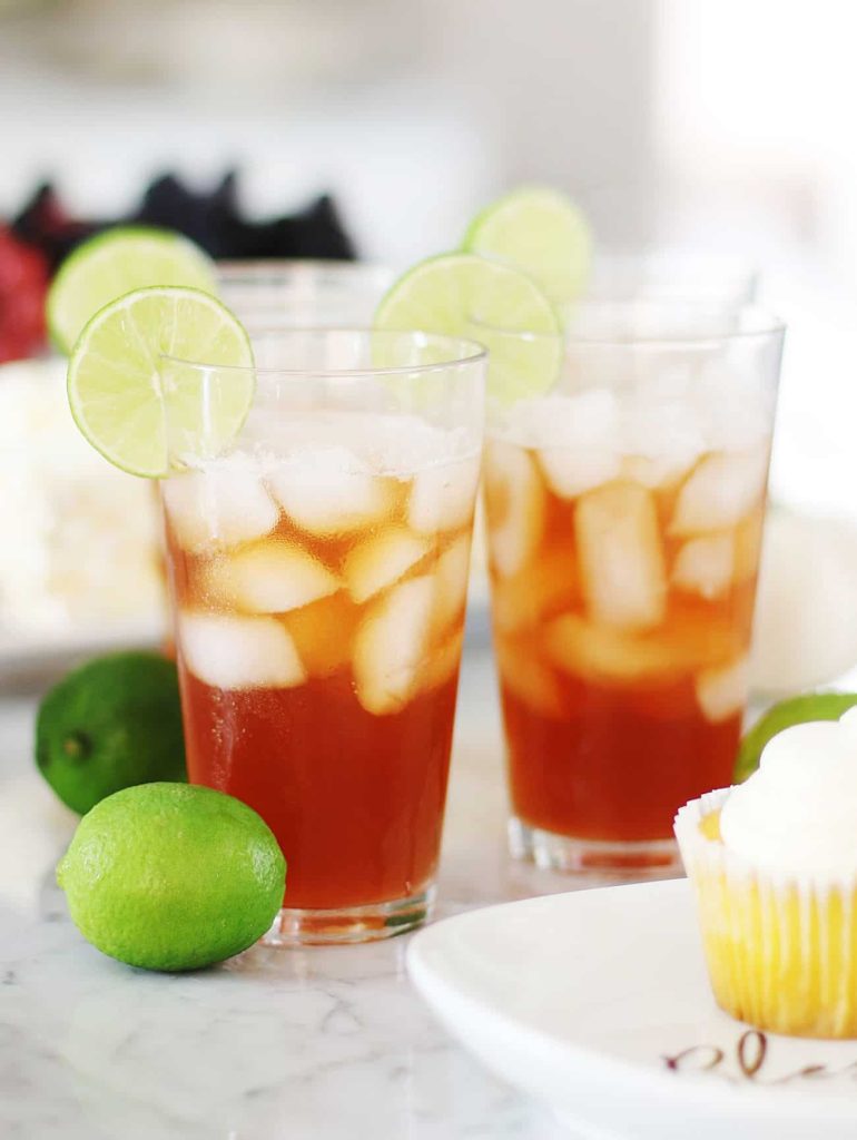 Three iced tea glasses with lime slices on the rims rest on a table, embodying the essence of the best summer drinks. Whole limes sit nearby, while in the background, a cupcake on a plate and assorted fruit complete this refreshing scene.