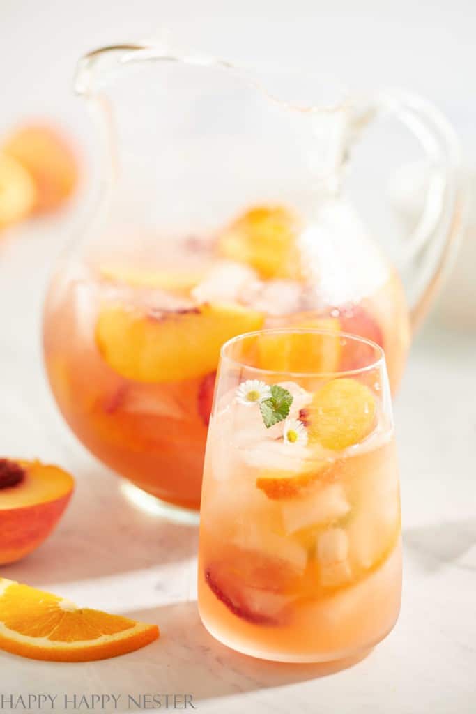 A glass and pitcher of peach sangria, one of the best summer drinks, sit on a table. Filled with peach slices and ice cubes, the glass is garnished with a small flower and mint leaf, while a sliced peach and orange wedge are nearby.