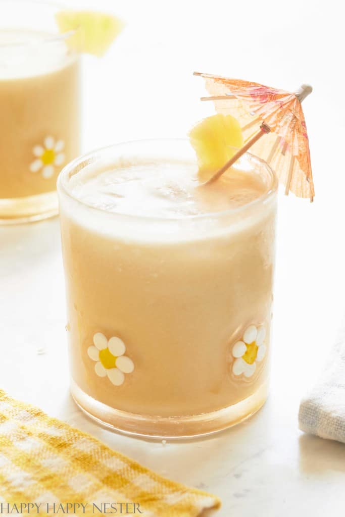 A creamy tropical drink, hailed as one of the best summer drinks, is served in a glass adorned with white and yellow flowers. The glass boasts a small paper umbrella and a pineapple wedge on the rim. Another similar concoction graces the background on a yellow checkered cloth.