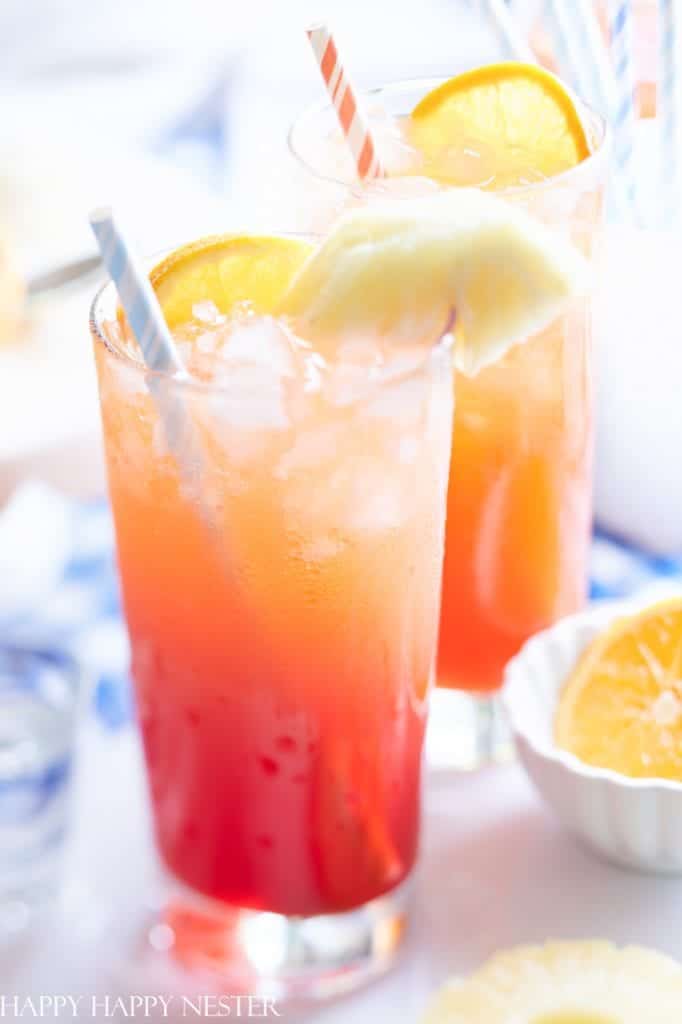 Two tall glasses of the best summer drinks, a vibrant orange and red layered delight, topped with ice. Each glass is garnished with a slice of orange and pineapple and includes a striped straw. Nearby, a bowl brims with fresh orange and pineapple slices.