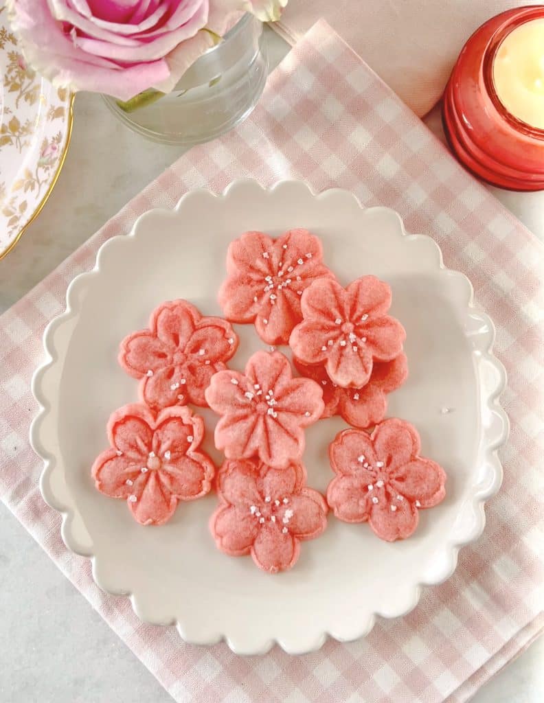 A white plate holds pink flower-shaped cookies, one of my favorite desserts, decorated with small white sugar pearls. The plate rests on a pink and white checkered napkin, beside a pink rose in a vase and a lit candle.