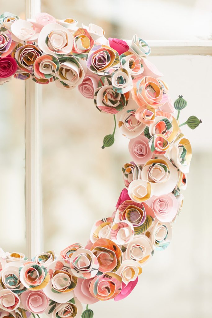 A decorative wreath made of colorful, paper-crafted roses with shades of pink, white, yellow, and green. Ideal for those who enjoy easy flower paper crafts, it hangs on a light-colored door or wall, showcasing intricate spiraled rose designs and green buds.