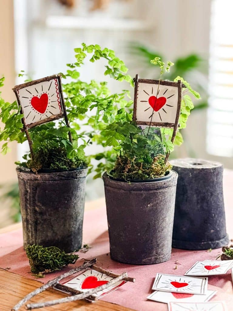 Two small potted plants with heart-shaped drawings on sticks grace the table, surrounded by candles adorned with dried flowers and additional heart drawings scattered nearby. The indoor setting is bathed in soft, natural lighting, creating a cozy atmosphere.