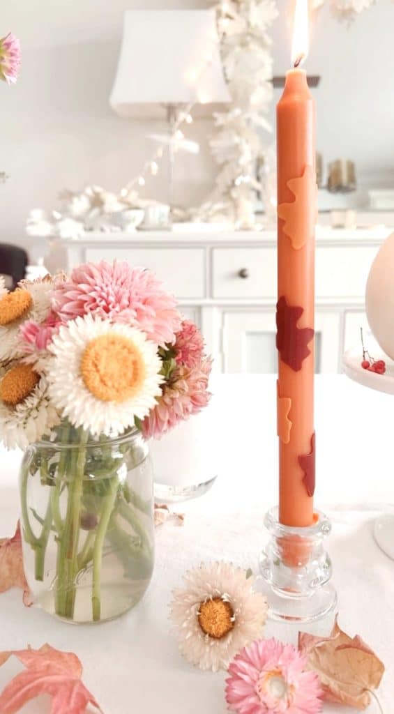 A tall orange candle with leaf decorations stands on a glass holder, embodying one of my favorite spring crafts. Next to it, a jar holds pink and white flowers with yellow centers. The background features a white dresser and soft lighting, creating a cozy ambiance.