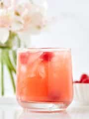 A glass filled with a pink, ice-cold drink garnished with cherries sits on a white surface, embodying one of those easy summer mocktail recipes. In the background, blurred white flowers and a small bowl of cherries complete the refreshing scene.