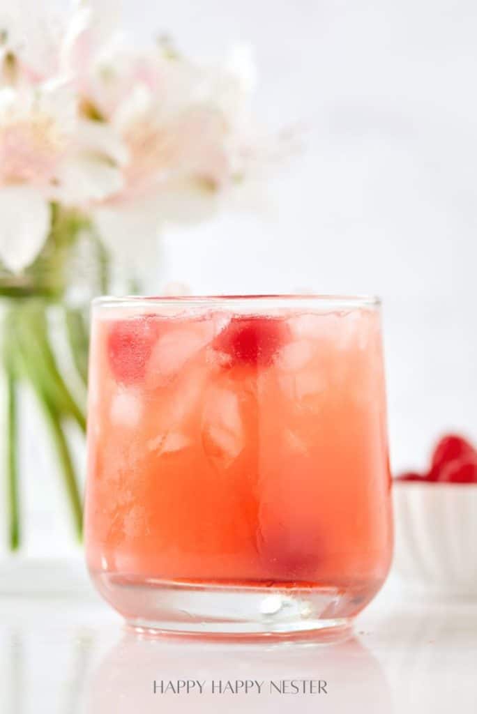 A glass filled with a pink, ice-cold drink garnished with cherries sits on a white surface, embodying one of those easy summer mocktail recipes. In the background, blurred white flowers and a small bowl of cherries complete the refreshing scene.