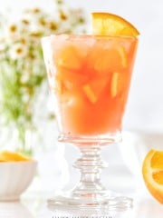 A glass of orange punch garnished with an orange slice, filled with ice and showing bits of orange pulp. In the background, white flowers in soft focus and an orange wedge in a small bowl are visible.