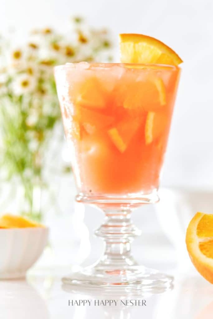 A glass of orange punch garnished with an orange slice, filled with ice and showing bits of orange pulp. In the background, white flowers in soft focus and an orange wedge in a small bowl are visible.