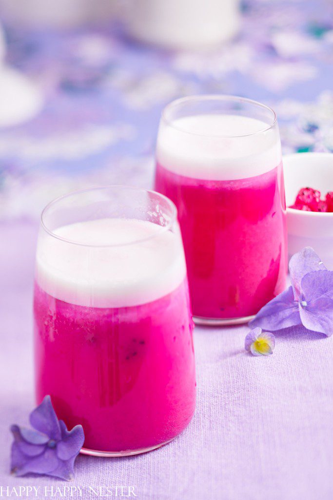 Two glasses of vibrant pink dragon fruit smoothie, among the best summer drinks, are topped with white froth on a pastel purple surface. Blue and purple flowers are scattered around, crafting a fresh and colorful setting.