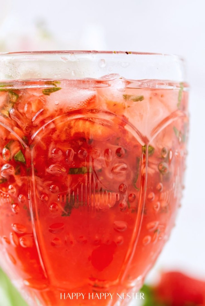 Close-up of a glass filled with a refreshing red Strawberry Basil Cocktail, garnished with fresh strawberries and mint leaves. The embossed patterns on the glass catch the light, while ice cubes glisten within. The softly blurred background adds an elegant touch to this delightful recipe.