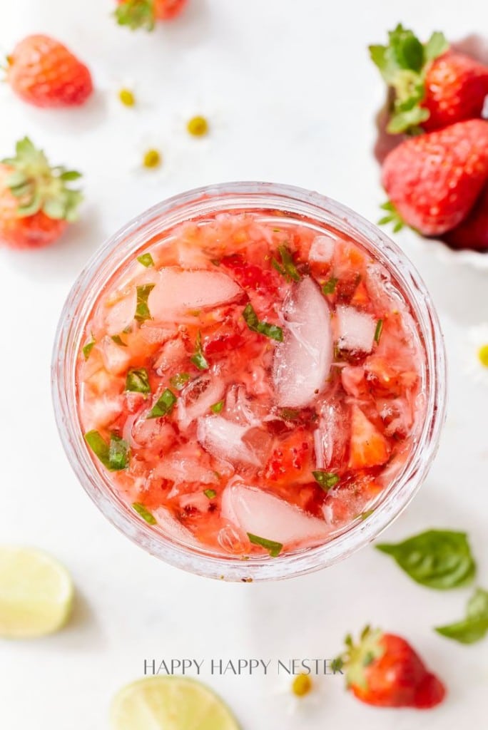 A refreshing strawberry basil cocktail with ice cubes, garnished with mint leaves in a glass. Surrounding the drink are fresh strawberries, lime wedges, and small white flowers on a white surface.