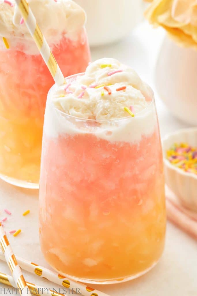 A colorful drink in a glass with crushed ice and layers of pink and orange hues. Topped with a scoop of ice cream and sprinkled with bits of pink and yellow sprinkles, this is one of the best summer drinks. A striped straw graces the glass, while another similar drink lingers enticingly in the background.