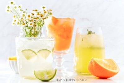 Three refreshing drinks are displayed: an easy summer mocktail with lime slices, an orange drink with ice, and a yellow drink with mint. A small bowl of orange wedges and a bouquet of daisies grace the marble surface in the background.