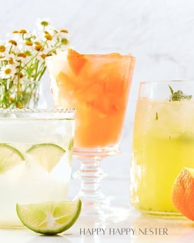 Three refreshing drinks are displayed: an easy summer mocktail with lime slices, an orange drink with ice, and a yellow drink with mint. A small bowl of orange wedges and a bouquet of daisies grace the marble surface in the background.