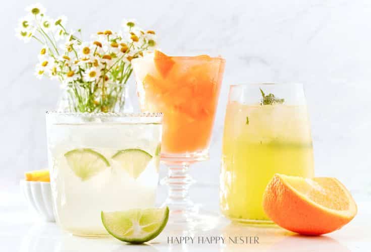 Three refreshing drinks are displayed: an easy summer mocktail with lime slices, an orange drink with ice, and a yellow drink with mint. A small bowl of orange wedges and a bouquet of daisies grace the marble surface in the background.