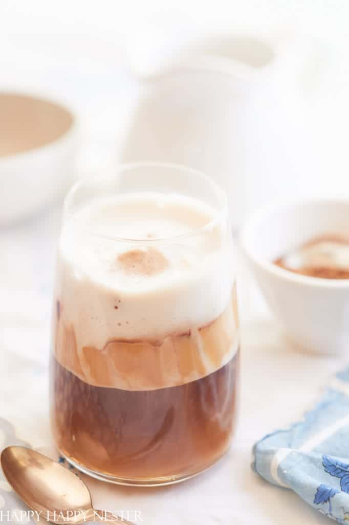 A glass of Dalgona coffee, one of the best summer drinks, sits on a table showcasing layers of frothy cream and dark coffee. Nearby, a spoon and blue-patterned napkin add charm, while a bowl and jug softly blur in the background.