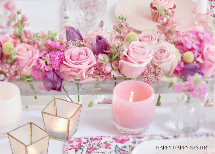 A Valentine’s table centerpiece featuring pink roses, purple flowers, and greenery in a rectangular glass vase is a romantic touch. Nearby, two lit candles in gold holders illuminate a pink cupcake on a white plate. A floral tablecloth adds charm to this enchanting setting.