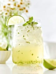 A refreshing mojito in a clear glass garnished with a lime slice and mint leaves. Ice cubes fill the glass, with herbs visible in the drink. A bowl of limes and a vase of daisies are blurred in the background.