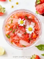 A refreshing strawberry basil cocktail with crushed strawberries, ice, and garnished with two small white flowers. Surrounded by fresh strawberries, lime slices, and basil leaves on a light background.